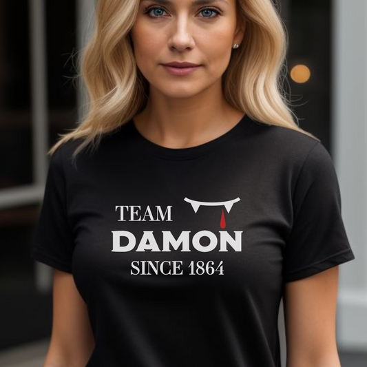 Young woman with long blonde hair wearing a black The Vampire Diaries Team Damon Since 1864 shirt with fangs and blood drip created by apparel shop Midnight Gypsy Designs.