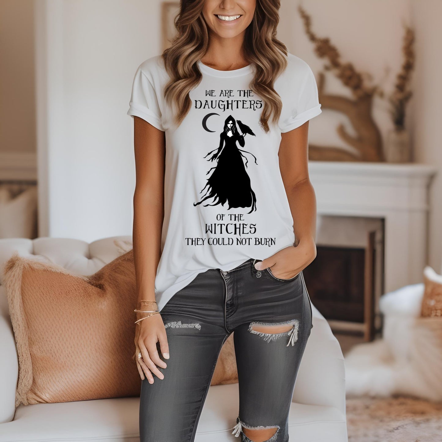 Young woman with long light brown hair smiling with hand in her pocket wearing grey jeans and a white t-shirt with black lettering that reads We Are the Daughters of the Witches They Could Not Burn with the moon and a witch in long black gown with a crow on her arm from apparel shop Midnight Gypsy Designs.