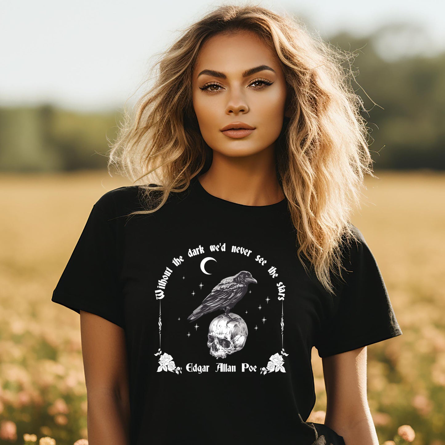 Young woman with long wavy blonde hair standing in a field of flowers wearing a black t-shirt with the saying Without the dark we'd never see the stars by Edgar Allan Poe with a crescent moon, stars, and a raven standing on a skull with roses created by apparel shop Midnight Gypsy Designs.